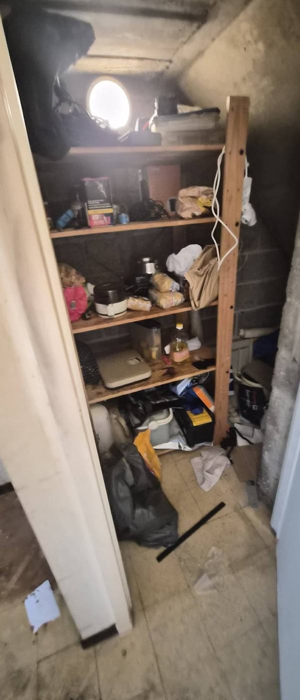 Donkere berging met volle houten rekken, dozen, keukentoestellen en een vuilzak op de betonvloer
