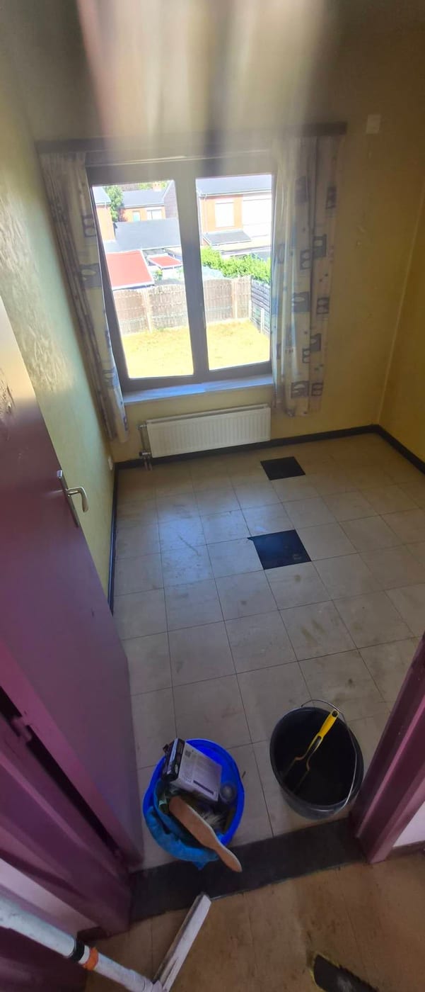 Dezelfde kamer leeggehaald, met dezelfde bedrukte gordijnen, de radiator weer vrij onder het raam en een gedweilde tegelvloer