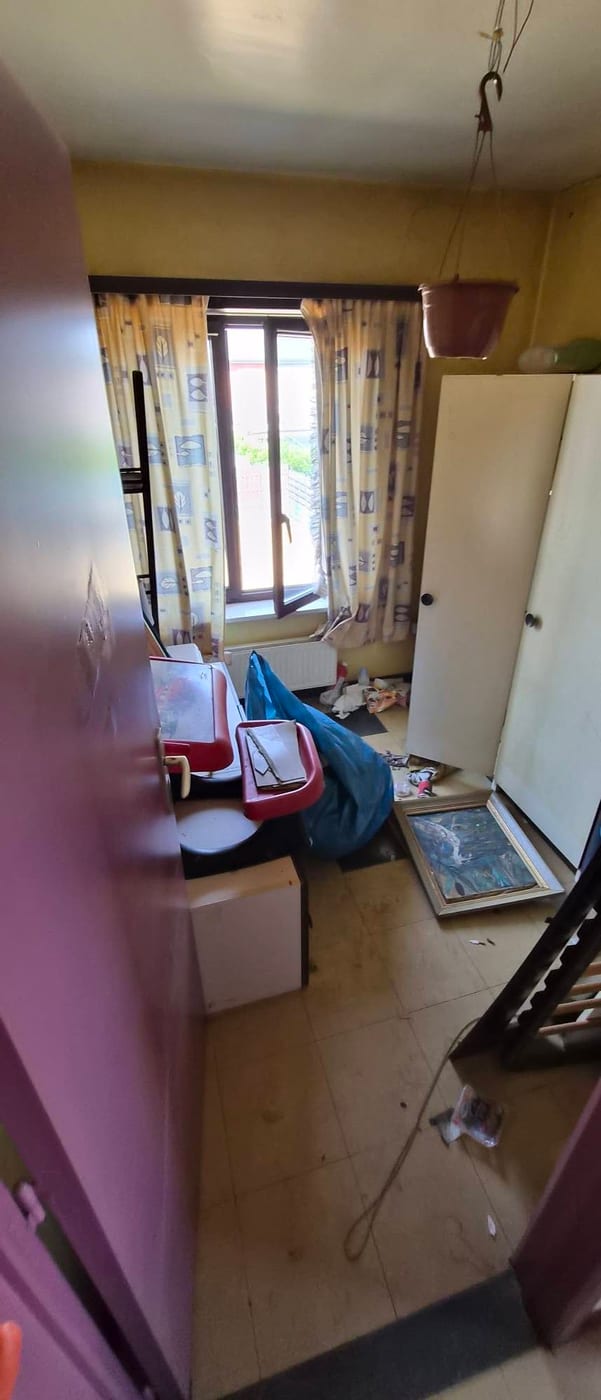 Kamer met okergele muur en bedrukte gordijnen, vol losse spullen, een losgeschroefde deur en een kinderstoeltje