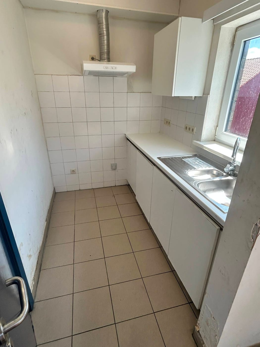 Kleine keuken met leeggehaalde kasten, een dubbele spoelbak onder het raam en een dampkap tegen de zijmuur