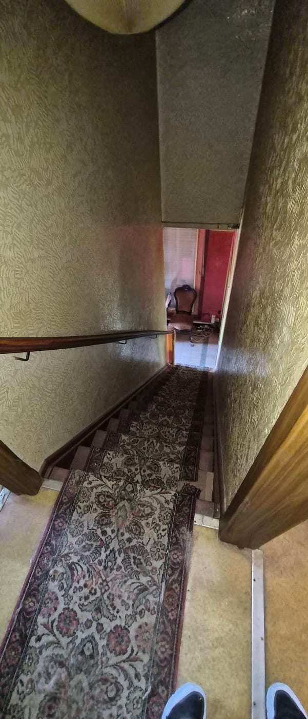 Steile trap met een versleten loper en gebloemd behang, met zicht op de kamer beneden